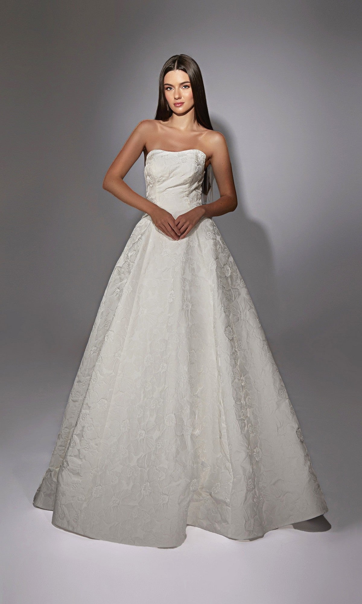 A woman stands against a plain background wearing the Alyce 7155 Long White Dress—a strapless, floor-length gown with a full skirt and subtle floral jacquard fabric.